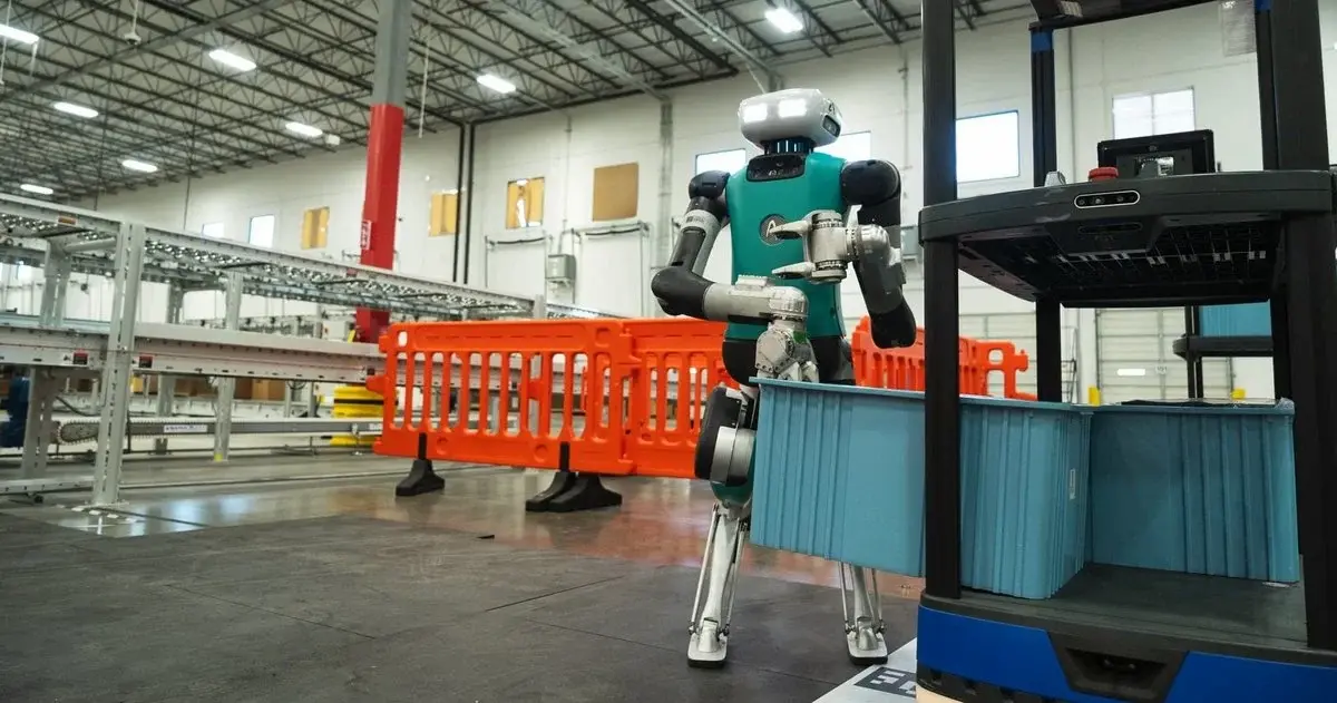 Góc nhìn rộng của một kho bãi logistics hiện đại, nơi robot hình người Digit đang giữ một thùng hàng màu xanh lam, với băng tải và rào cản màu cam ở hậu cảnh.