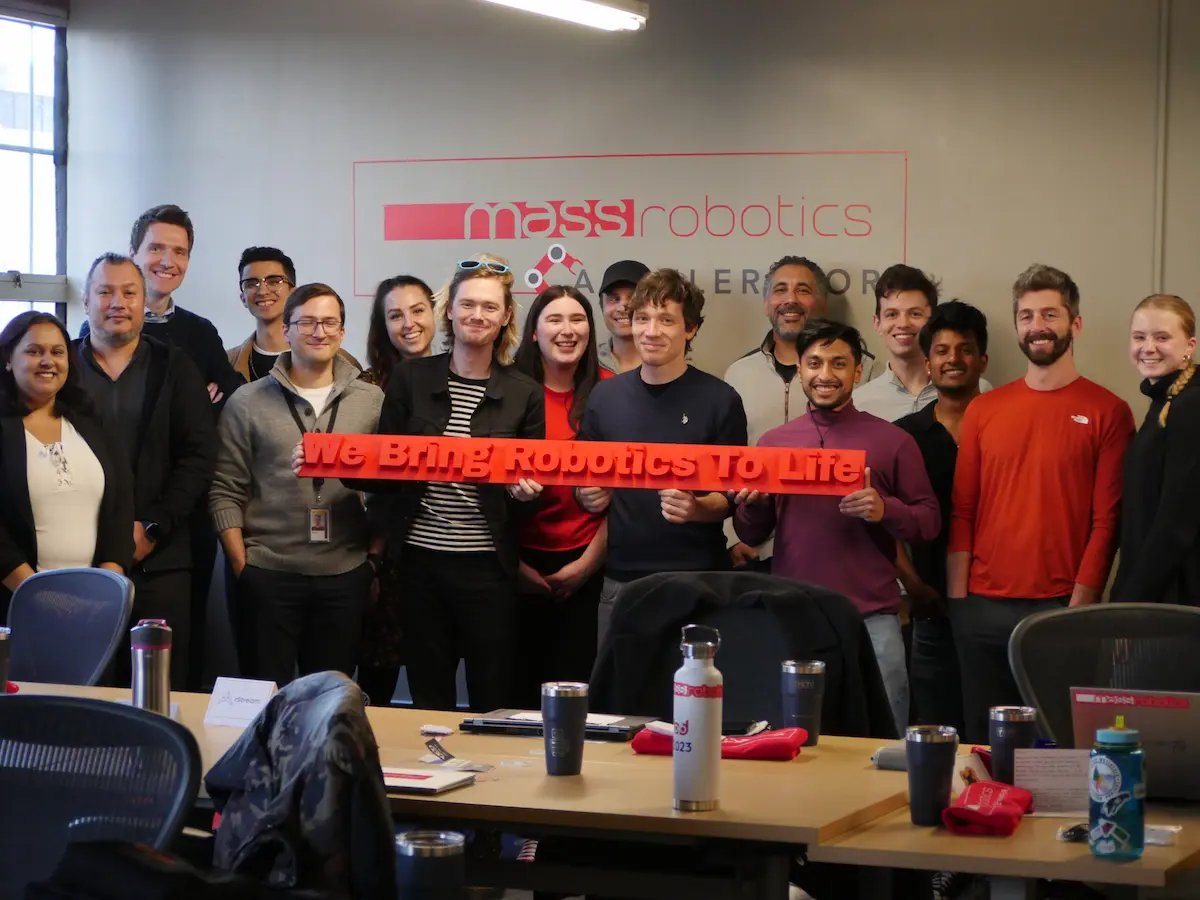Một nhóm người tại MassRobotics chụp ảnh tập thể cầm biển hiệu "We Bring Robotics To Life".