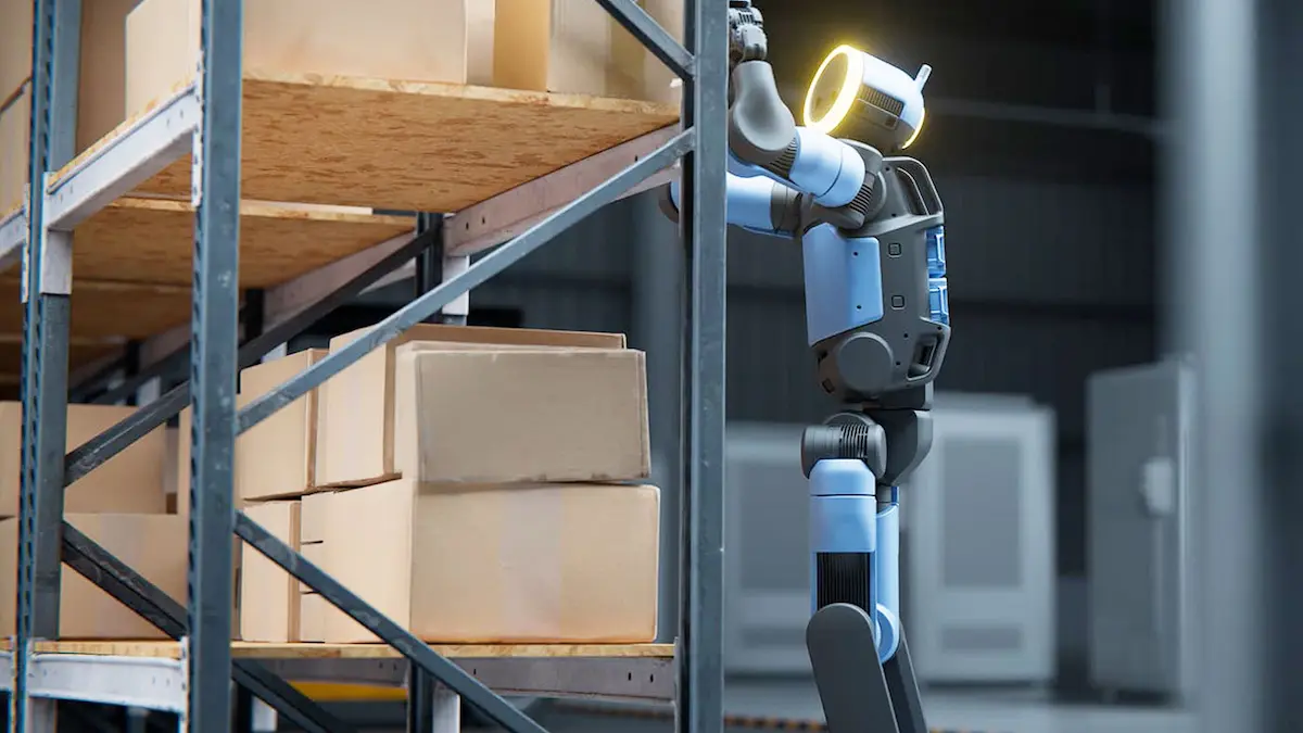 Robot hình người Atlas thuần điện thế hệ mới đứng và cầm một hộp các tông bằng cả hai tay cạnh kệ hàng.