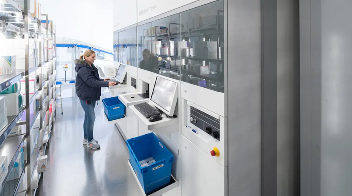 Phụ nữ làm việc tại trạm làm việc của hệ thống tự động hóa nhà thuốc BD Rowa Vmax, dường như nạp thuốc cho robot (Bản thu nhỏ)